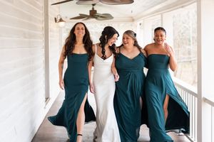 Bride in white gown walks with three bridesmaids in teal dresses on a porch, laughing and celebrating together.