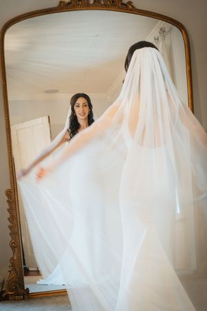 Bride in a flowing white gown and veil admires her reflection in an ornate gold-framed mirror before her wedding.
