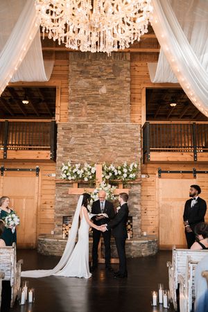 Couple exchanges vows beneath a grand chandelier in a rustic barn venue with stone fireplace and white florals.