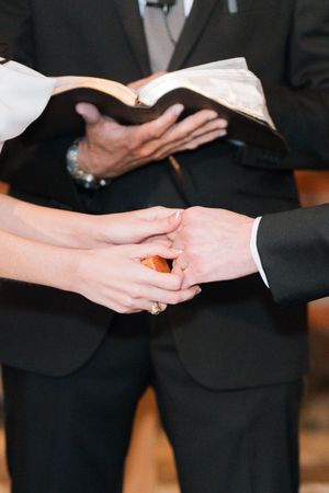 Couple holding hands during wedding ceremony with officiant holding Bible in background.