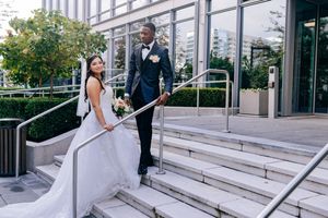 A bride and groom pose on modern building steps with glass walls and greenery in the background.