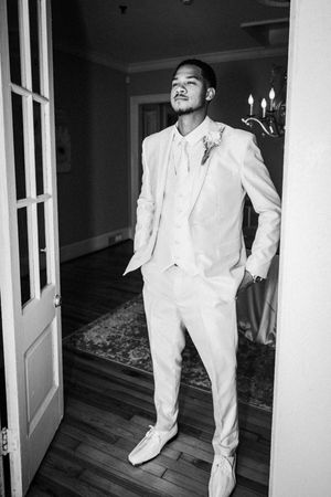 Elegant groom in white suit posing confidently in doorway during wedding day preparations.