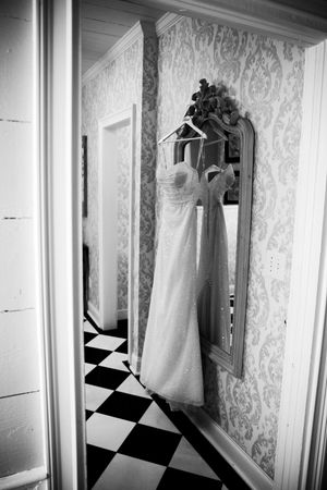 A white wedding dress hangs on an ornate mirror in a hallway with black and white checkered floors.