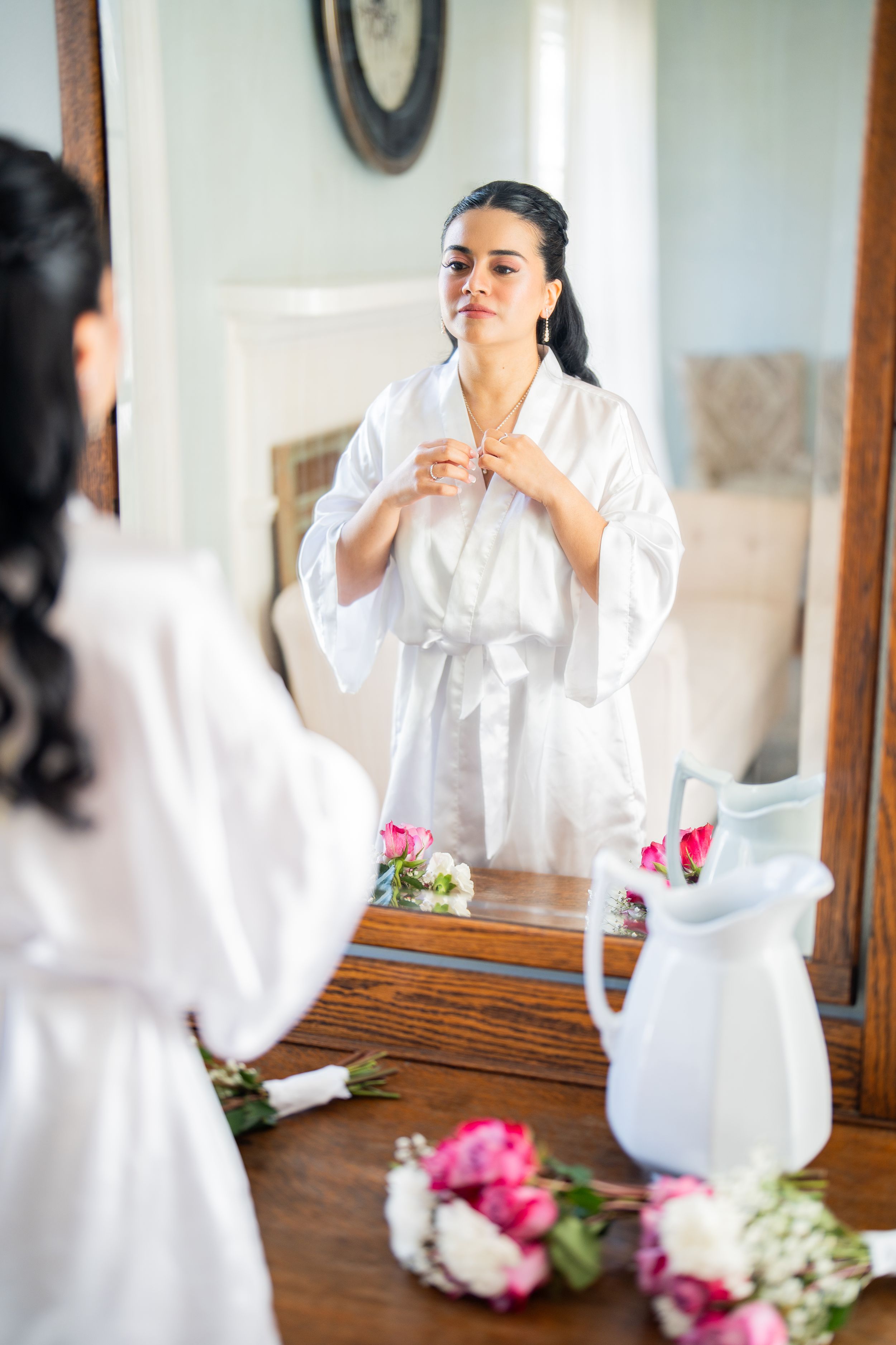 Person in white robe getting ready at vanity with pink flowers and white vase on wooden table.