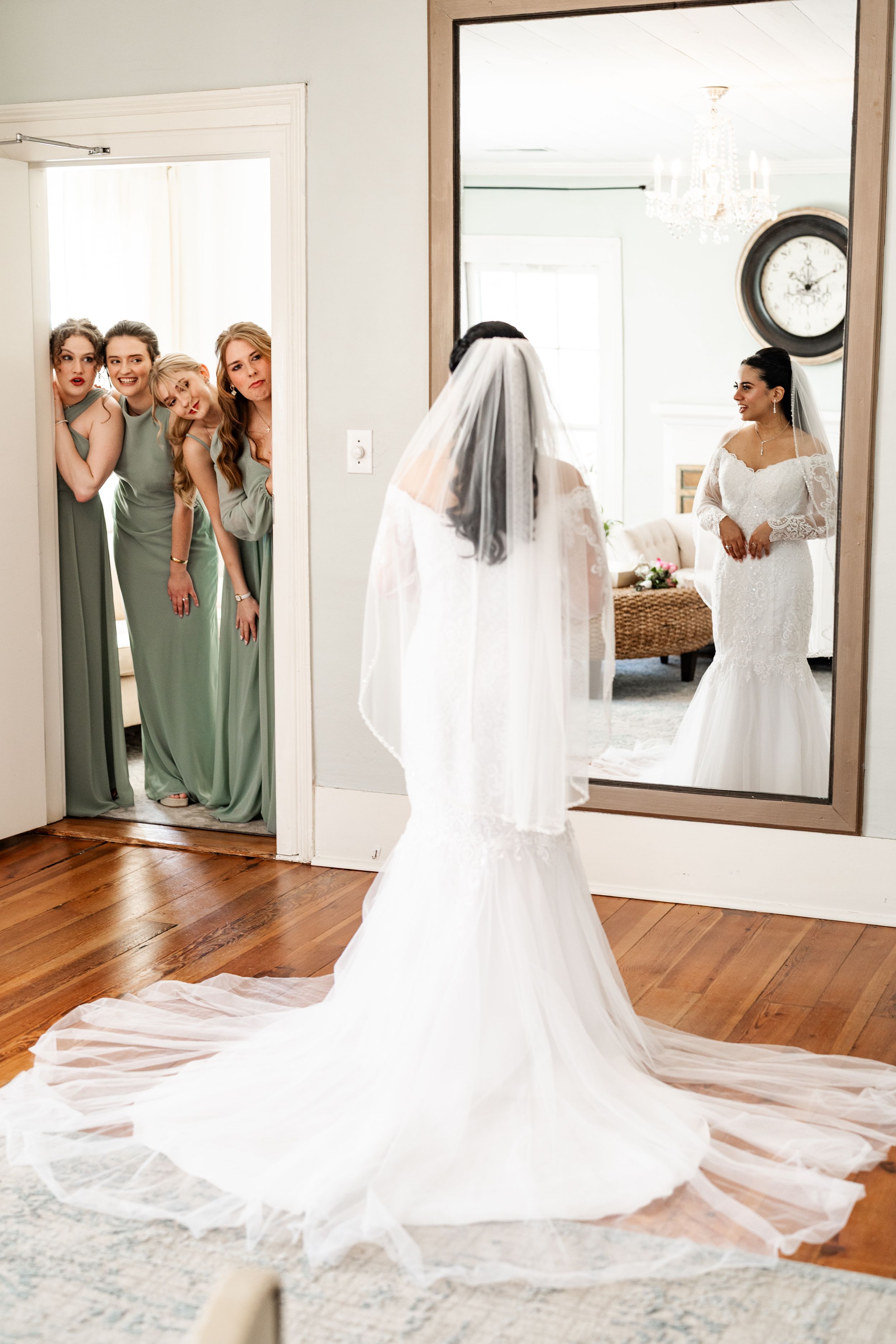 A wedding dress with a long flowing train is displayed in a bridal suite while bridesmaids peek through the doorway.