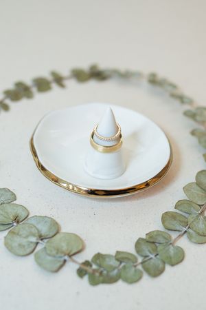 Gold wedding rings on white ceramic ring holder surrounded by eucalyptus leaves wreath on light background.