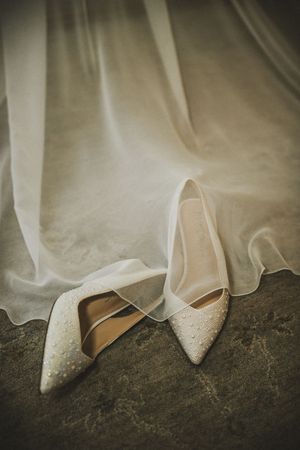 Pearl-embellished bridal heels resting on stone surface beneath a flowing white wedding veil.