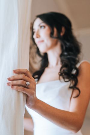 Close-up of bride wearing elegant halo diamond engagement ring while holding sheer curtain near window.