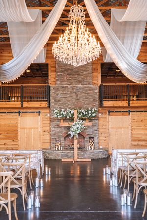 Elegant barn wedding ceremony space with crystal chandelier, white draping, and stone fireplace altar.
