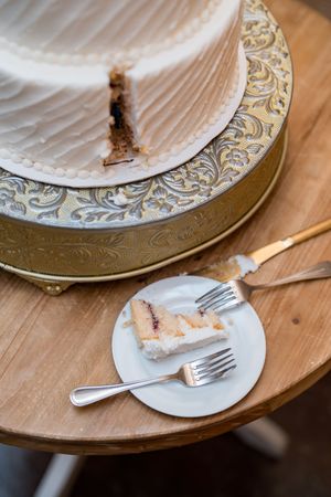 Two-tier white wedding cake on ornate gold stand with sliced piece on small plate.