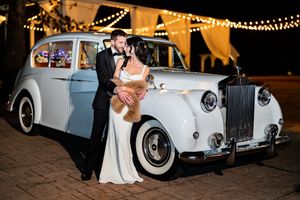 Bride and groom pose elegantly beside a classic white Rolls-Royce under twinkling string lights.