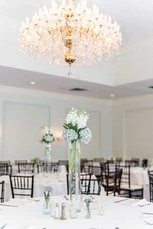 Tall centerpiece with white hydrangeas in a crystal vase under an ornate chandelier.