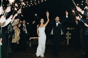 Bride and groom exit wedding reception through sparkler tunnel at night, celebrating with raised hands.
