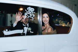 Smiling newlyweds wave from a white Just Married car window after their wedding.