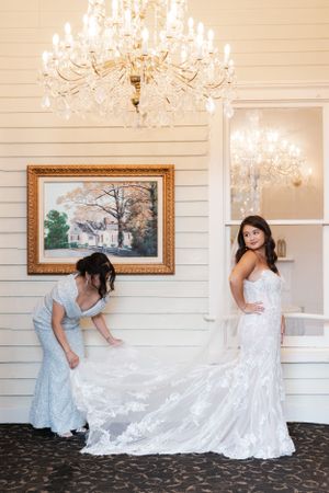 Elegant wedding gown with intricate lace detailing being arranged in a room with crystal chandeliers and artwork.