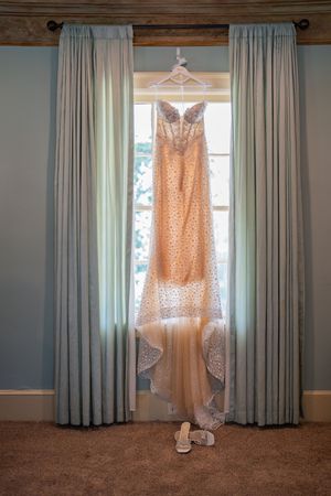 Elegant lace wedding dress hanging in a sunlit window with flowing train and bridal shoes below.