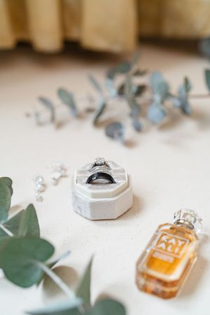 Wedding rings in white hexagonal box with diamond engagement ring, Kayali perfume and eucalyptus greenery.