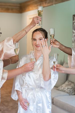 Bride in white satin robe shows off engagement ring while bridesmaids raise champagne glasses in celebration.