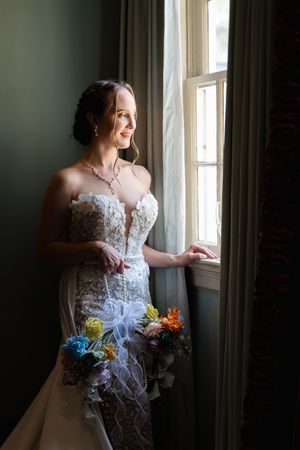 Radiant bride in lace gown holds colorful bouquet while gazing out sunlit window.