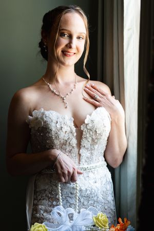 Radiant bride in strapless lace gown with pearl necklace smiling by window on wedding day.