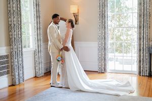 Interracial bride and groom sharing romantic moment in elegant room, bride in white gown, groom in cream suit.