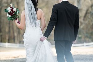 A couple in wedding attire walks through a wooded area holding hands and carrying floral bouquets.