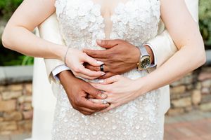 Close-up of interracial couple's hands with wedding rings, bride in floral lace gown, groom wearing a watch.