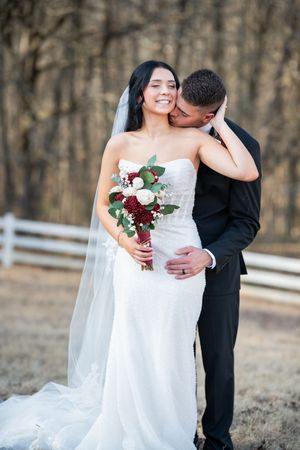 A romantic wedding couple embraces outdoors in winter with the bride holding a burgundy and white bouquet.