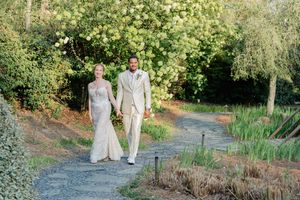 Newlywed couple walks hand in hand along garden path surrounded by blooming trees.