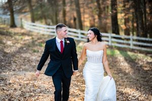 A couple in wedding attire walks along a rustic fence in an autumn forest setting during their fall wedding celebration.
