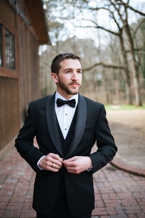 Groom in black tuxedo with bow tie buttoning jacket outdoors near rustic wooden building.