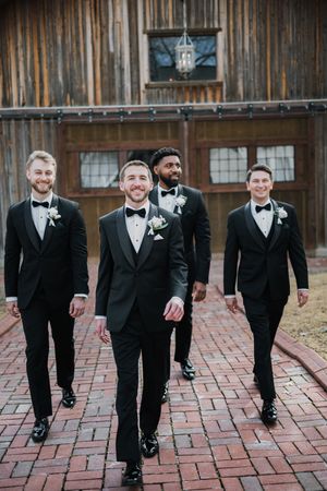 Groom and three groomsmen in black tuxedos walk confidently outside a rustic barn.
