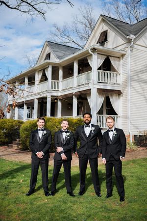 Groom and three groomsmen in matching black tuxedos pose outdoors in front of a white historic venue.