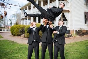 Groomsmen toss smiling groom into the air outside a white wedding venue.
