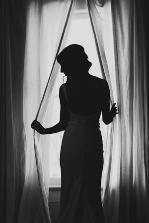 Elegant black and white silhouette of bride in wedding gown standing between sheer curtains at a window.