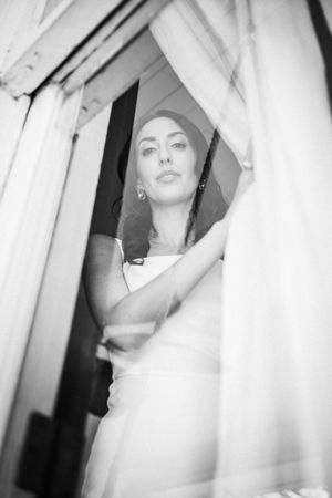 Elegant bride in white dress gazing through sheer curtains in black and white photo.
