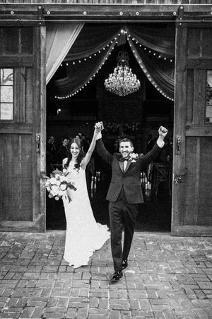 Black and white photo of newlyweds raising hands exiting a barn with chandelier and string lights.