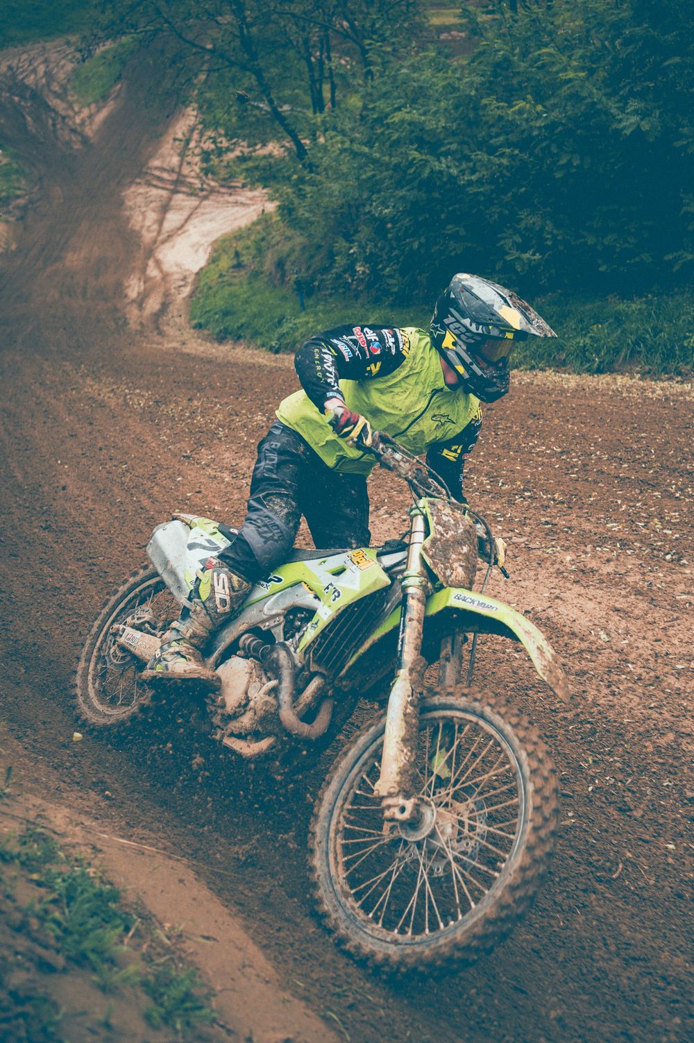 Motocross rider in neon yellow gear and helmet leans a lime green dirt bike through a muddy, rutted forest trail.