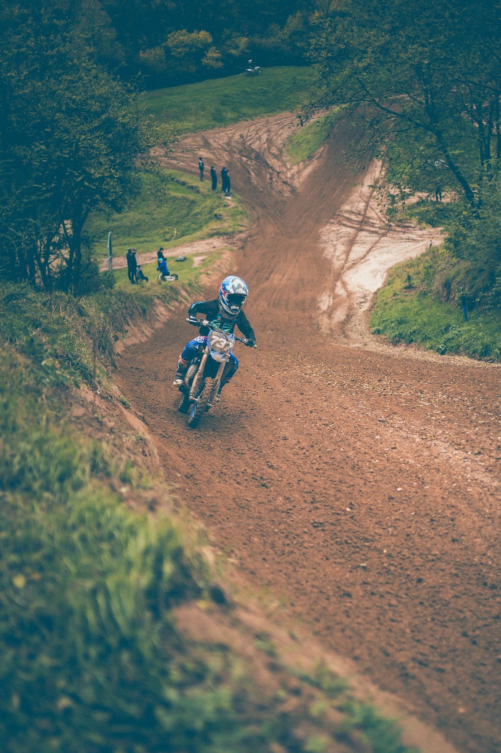 A motocross rider in a blue helmet races a dirt bike down a muddy hillside track with spectators and green trees in the background.