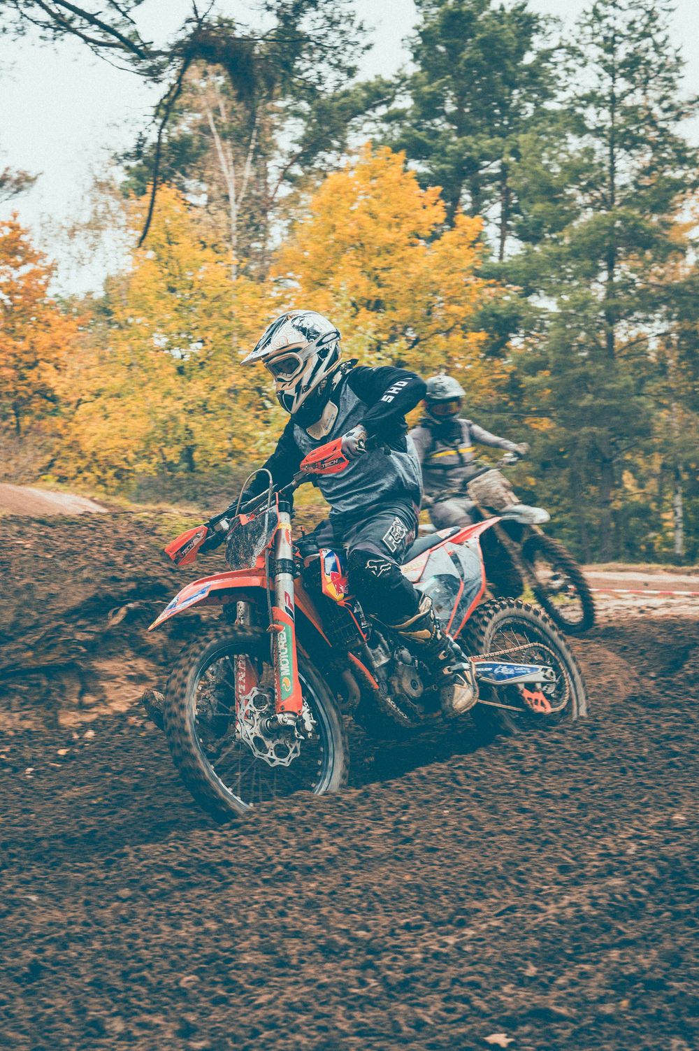 Motocross rider in a white helmet and protective gear races an orange dirt bike over a muddy track with autumn trees in the background.