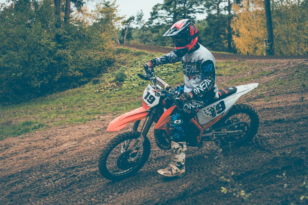 Motocross rider in a red helmet and racing gear riding an orange dirt bike on a muddy forest trail surrounded by autumn trees.