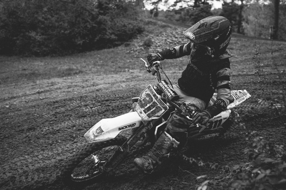 Black and white photo of a helmeted dirt bike rider leaning into a muddy corner on a motocross track surrounded by trees.