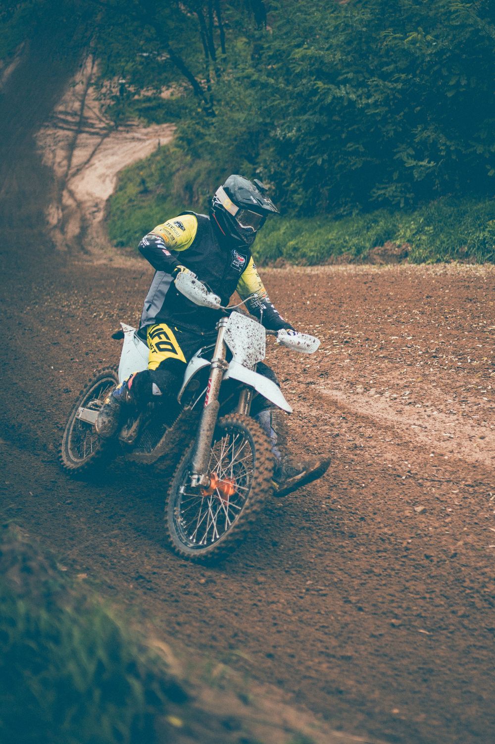 Motocross rider in yellow and black gear leans a white dirt bike into a turn on a muddy off-road trail surrounded by green trees.