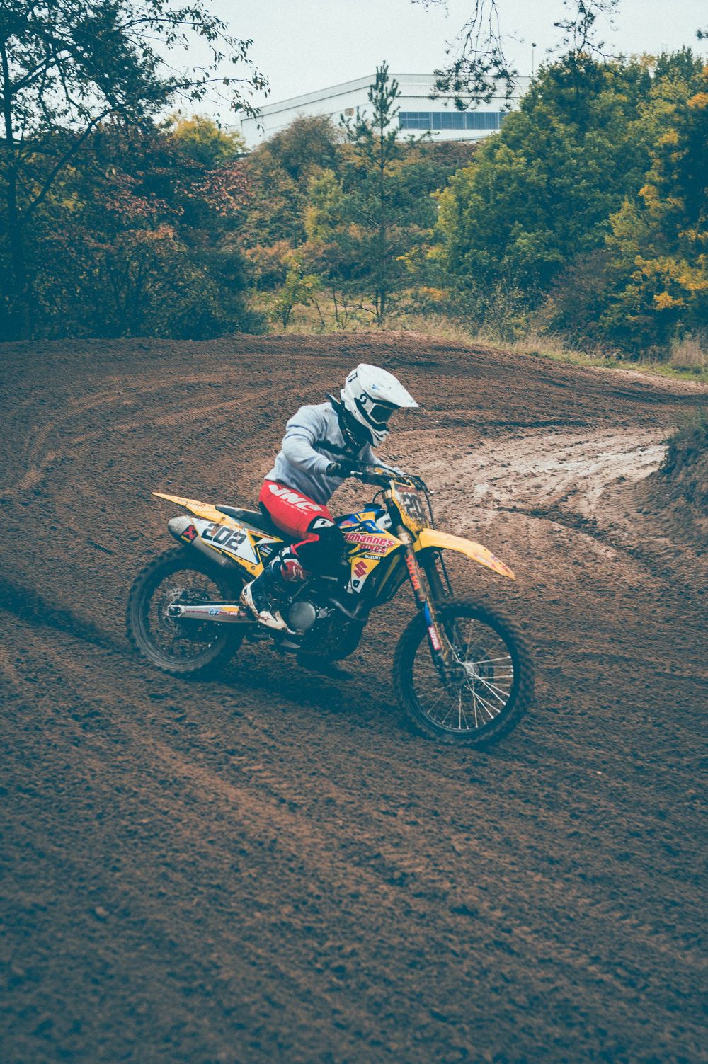 Motocross rider in a white helmet and red pants leans a yellow dirt bike through a muddy berm on an outdoor off-road track.