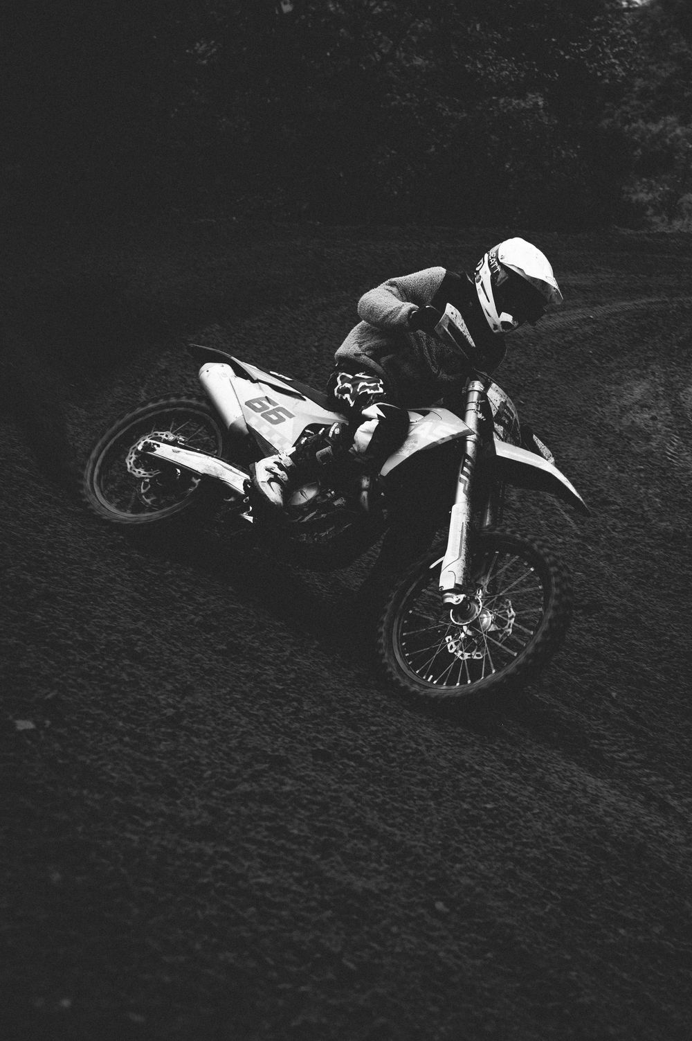 Black and white photo of a motocross rider in a helmet leaning a dirt bike into a sharp turn on a dark, rutted off-road track.