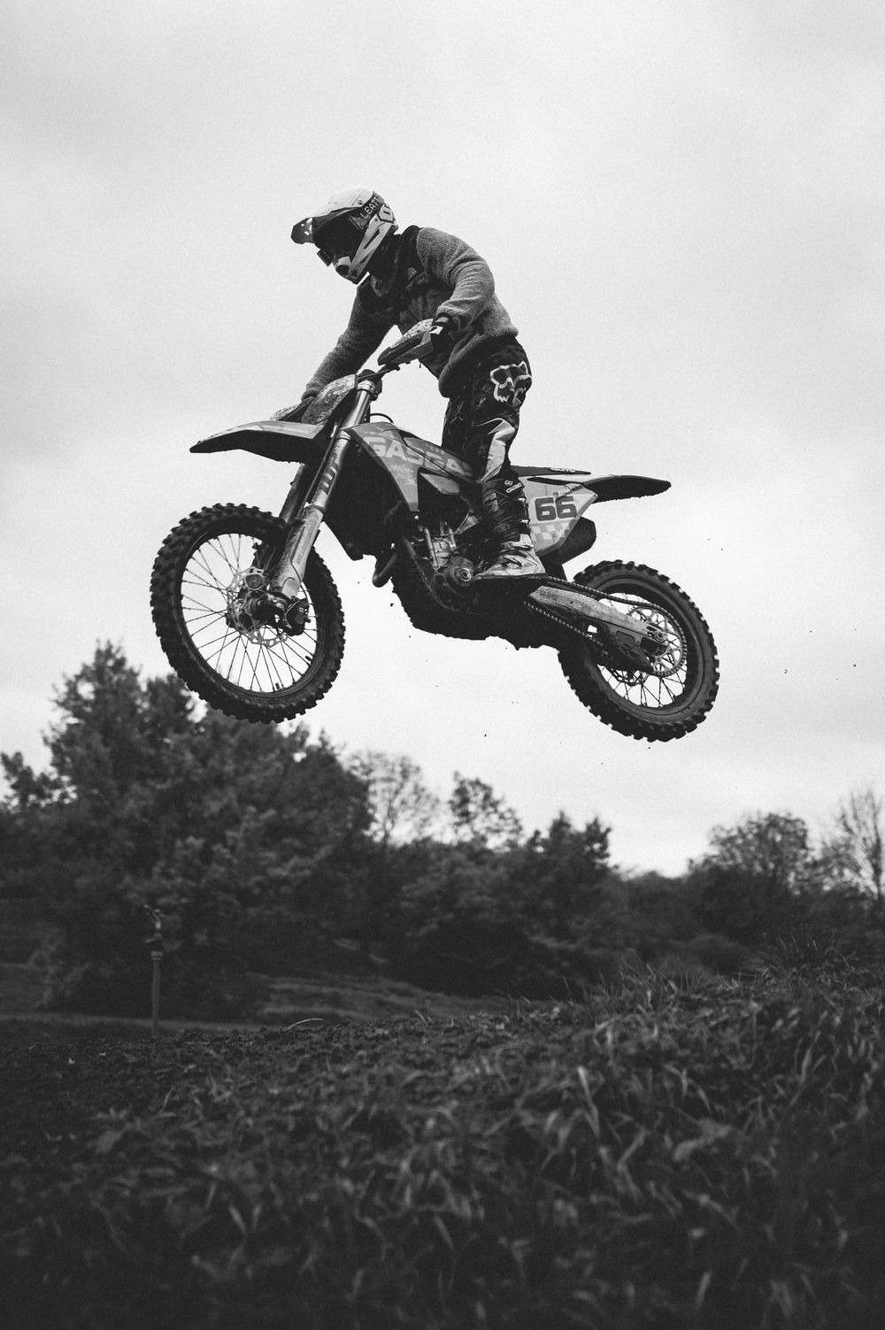 Black and white photo of a motocross rider in helmet and gear launching a dirt bike through the air above a grassy track with trees behind.