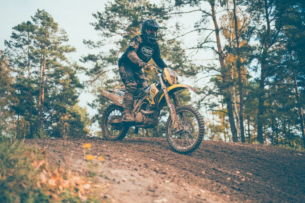 Motocross rider in a helmet and riding gear races a dirt bike up a sandy forest trail surrounded by tall pine trees.
