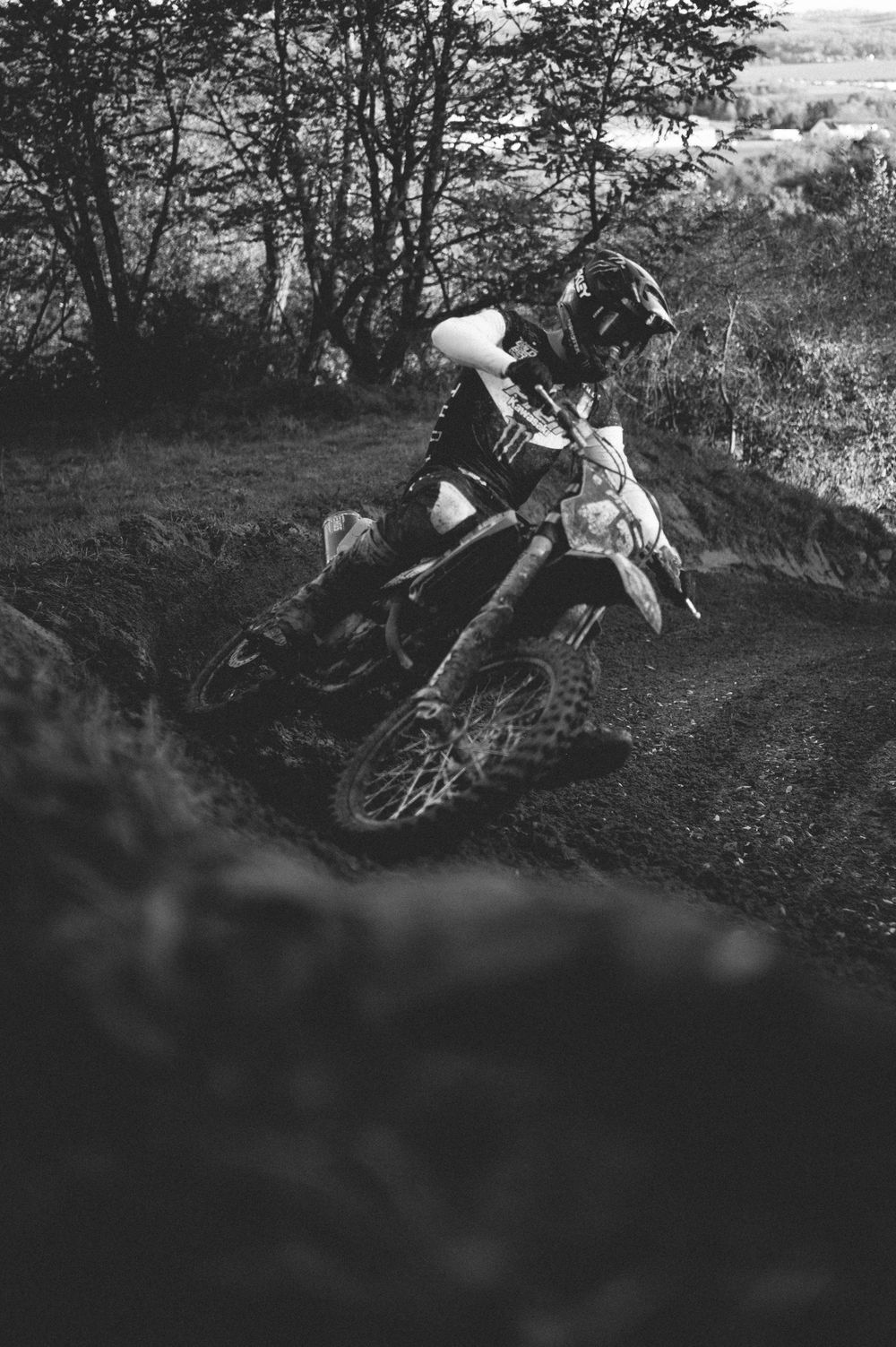 Black and white photo of a motocross rider leaning into a sharp turn on a muddy dirt track surrounded by trees.