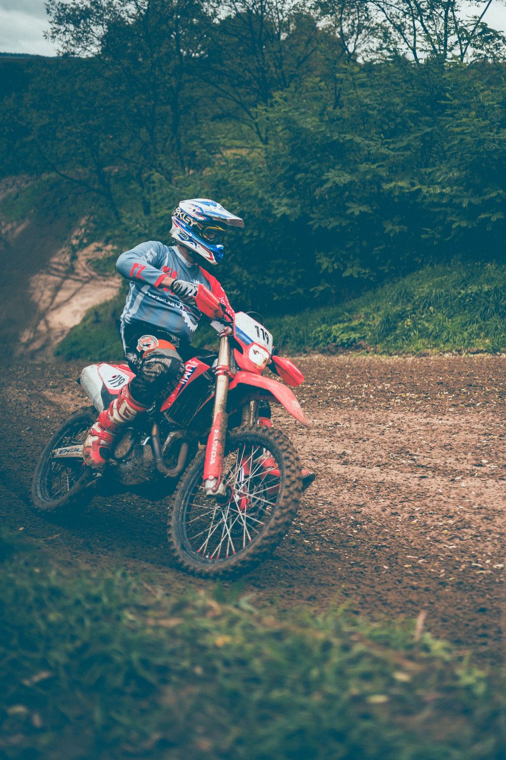 A motocross rider in a blue helmet and gear leans a red dirt bike through a muddy turn on a forest track surrounded by green trees.