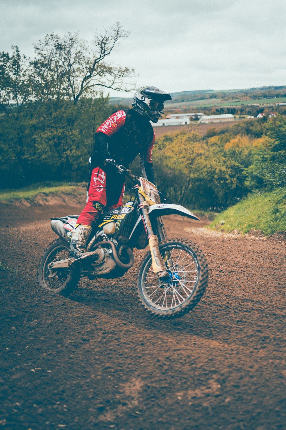 Motocross rider in red gear and helmet rides a dirt bike over a muddy track with green hills and trees in the background.